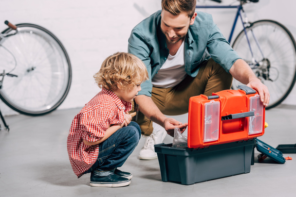 father and adorable little son sitting near opened tools box in bicycle workshop - Foto, imagen