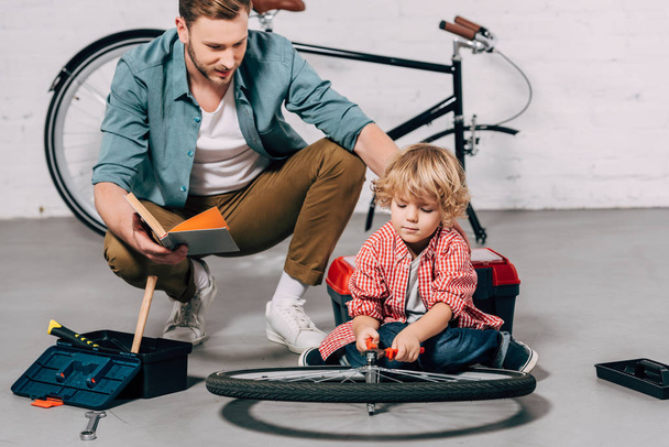 селективный фокус человека с книгой, сидящего рядом с маленьким сыном, фиксирующим велосипедное колесо плоскогубцами в мастерской
 - Фото, изображение
