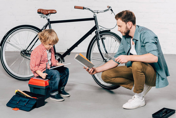 dziecko siedzi z cyfrowego tabletu podczas ojciec czytanie książki o rower warsztat - Zdjęcie, obraz