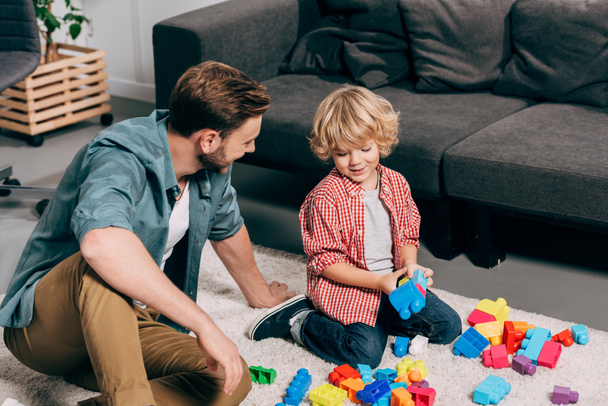 high angle view of happy child and his father playing with colorful plastic blocks on floor at home - Photo, Image