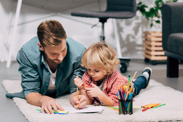 selective focus of father and son drawing on white paper by colorful markers at home - Фото, изображение