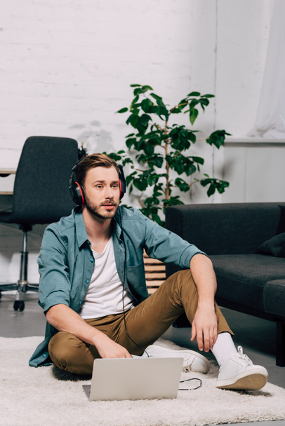 young male freelancer in headphones working on laptop while sitting on floor at home - 写真・画像