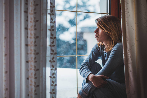 Λυπημένη γυναίκα που κάθεται δίπλα στο παράθυρο κοιτάζοντας έξω από - Φωτογραφία, εικόνα