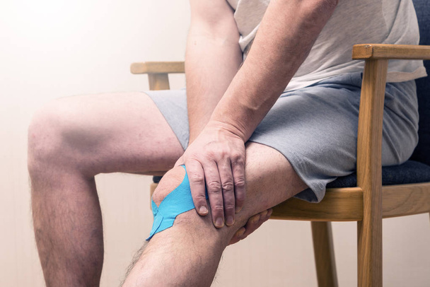 Man sitting on the chair and holding his sore knee with Kinesiol - Foto, Imagem