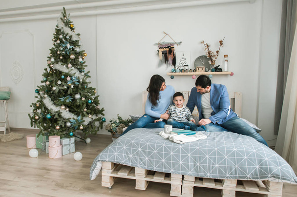 Young happy family of three laying on bed near christmas tree. Mother and father having fun with son in living room. Happy parenting concept. New Year's interior - Foto, afbeelding