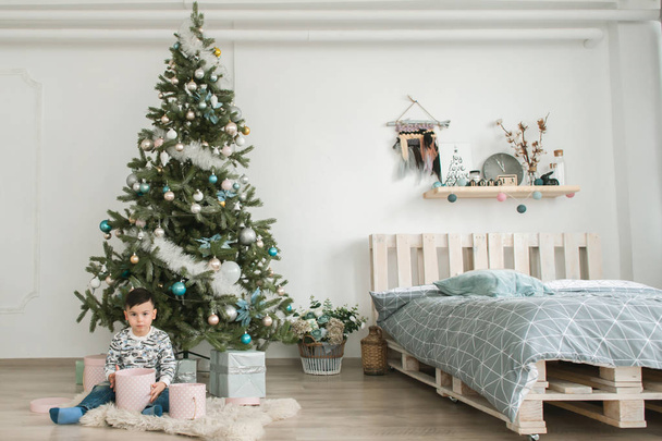 Little boy with gift box near Christmas tree on Christmas eve at home. Young kid in light bedroom with winter decoration. Happy family at home. Christmas New Year december time for celebration concept - Fotografie, Obrázek