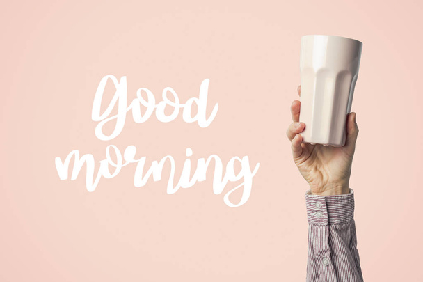 Female hand in clothes holding a purple cup with hot coffee or tea on a pink background. Added text Good morning. Breakfast concept with hot coffee or tea. - Foto, afbeelding