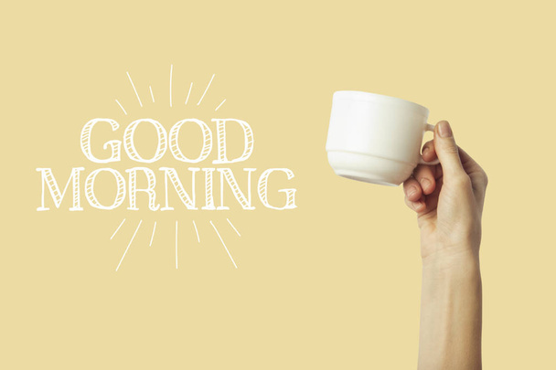 Female hand holding a green cup with hot coffee or tea on a light yellow background. Added text Good morning. Breakfast concept with hot coffee or tea. - Фото, изображение