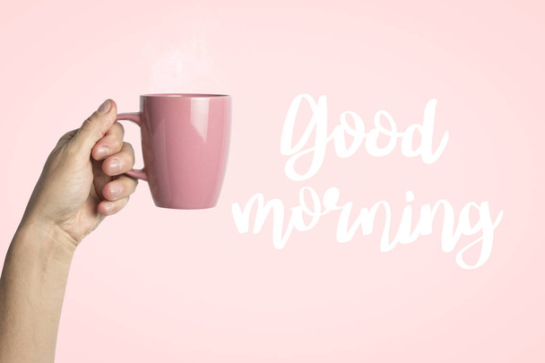 Vrouwelijke hand in kleren houden een paarse kop met warme koffie of thee op een roze achtergrond. Toegevoegde tekst Goedemorgen. Ontbijt concept met warme koffie of thee. - Foto, afbeelding