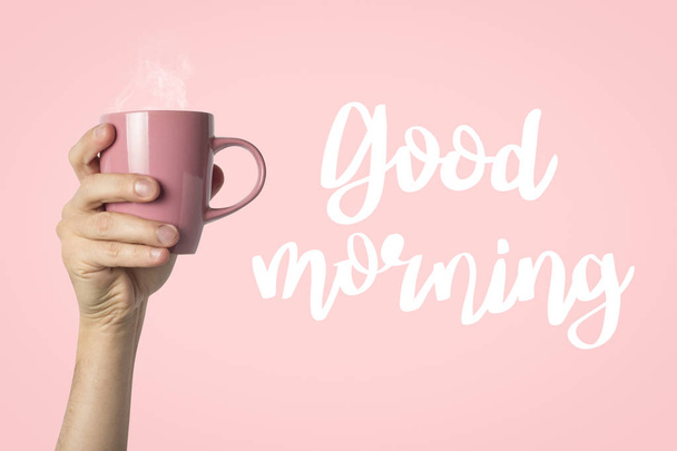 Female hand in clothes holding a purple cup with hot coffee or tea on a pink background. Added text Good morning. Breakfast concept with hot coffee or tea. - Foto, Bild