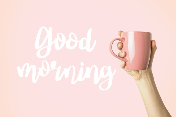 Mano femminile in vestiti che tengono una tazza viola con caffè caldo o tè su uno sfondo rosa. Aggiunto testo Buongiorno. Prima colazione con caffè caldo o tè
. - Foto, immagini