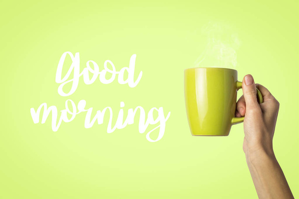 Mano femminile che tiene una tazza verde con caffè caldo o tè su uno sfondo verde chiaro. Aggiunto testo Buongiorno. Prima colazione con caffè caldo o tè
. - Foto, immagini