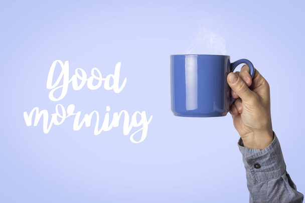 männliche Hand hält eine blaue Tasse mit heißem Kaffee oder Tee auf blauem Hintergrund. Text Guten Morgen. Frühstückskonzept mit heißem Kaffee. - Foto, Bild