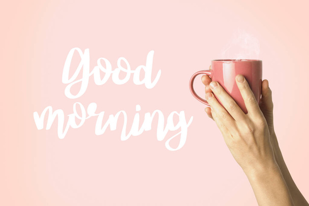 Nainen käsi vaatteita tilalla violetti kuppi kuumaa kahvia tai teetä vaaleanpunainen tausta. Lisätty teksti Hyvää huomenta. Aamiainen käsite kuumaa kahvia tai teetä
. - Valokuva, kuva