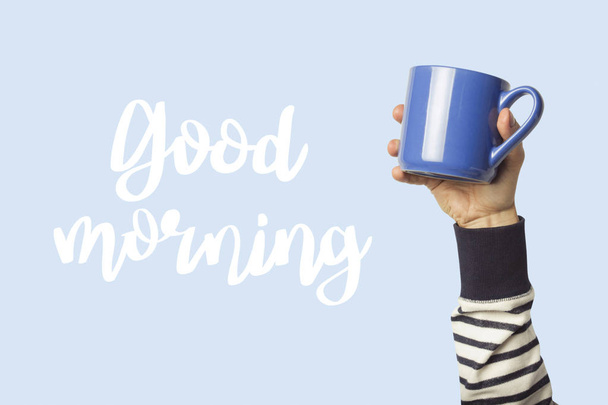 Male Hand is holding a blue cup with hot coffee or tea on a blue background. Added text Good morning. Breakfast concept with hot coffee. - Photo, Image