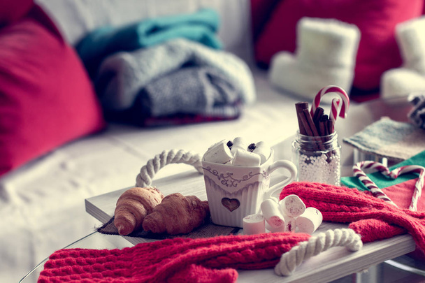 уютный зимний натюрморт, состоящий из чашки кофе, зефира, круассаны, полосатый леденец на переднем плане и меховые ботинки с свитерами в домашней гостиной
 - Фото, изображение