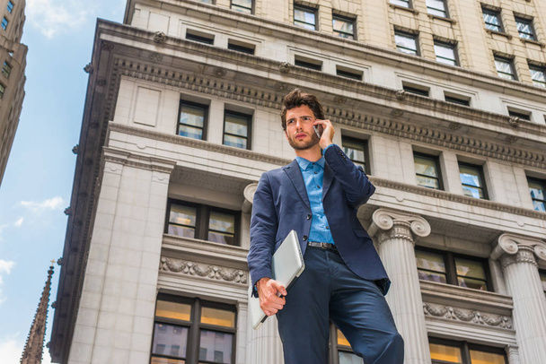 Сила технологии в нашей современной повседневной жизни. Одетый в синий костюм, молодой европейский бизнесмен с бородой, стоящий снаружи в Нью-Йорке, рука с ноутбуком, разговаривающий по мобильному телефону
 - Фото, изображение
