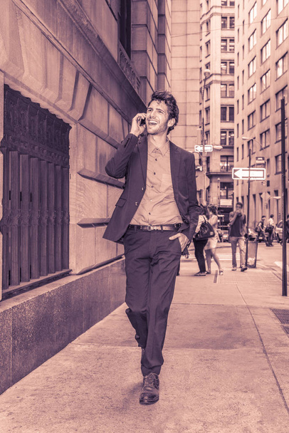 European businessman traveling in New York. Young guy with beard walking through crowded high building street, talking on mobile phone, smiling, excited with good news. Technology in Daily Life - Photo, Image