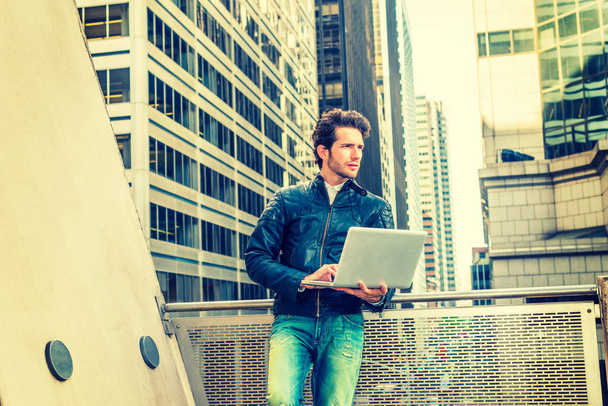 European businessman working in New York. Wearing black leather jacket, jeans, young guy with beard, standing by railing on balcony in business district, working on laptop computer. - Fotografie, Obrázek