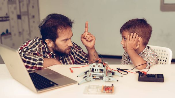 Bearded Father Help Upset Son with Robot at Home. Young Boy in Shirt. Using Laptop. Modern Hobby Concept. Modern Technology. Robot Engineering Concept. Bearded Young Man. Innovation for Fun. - Фото, зображення
