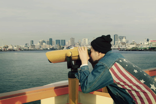 hipster man wearing american flag jeans jacket looking through binocular lens against urban building - Photo, Image