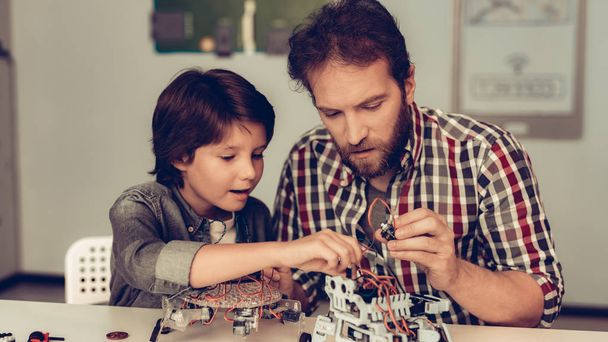 Bearded Father and Son Constructing Robot at Home. Young Boy in Shirt. Indoor Joy. Modern Hobby Concept. Modern Technology. Robot Engineering Concept. Bearded Young Man. Innovation for Fun. - Zdjęcie, obraz