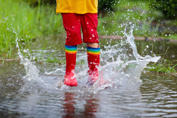 Dzieciak bawi się w deszczu. Dzieci w butach z parasolem i deszczem bawią się na świeżym powietrzu podczas ulewnego deszczu. Mały chłopiec skaczący w błoconej kałuży. Dzieci bawią się deszczową jesienną pogodą. Dziecko biegające w burzy tropikalnej. - Zdjęcie, obraz