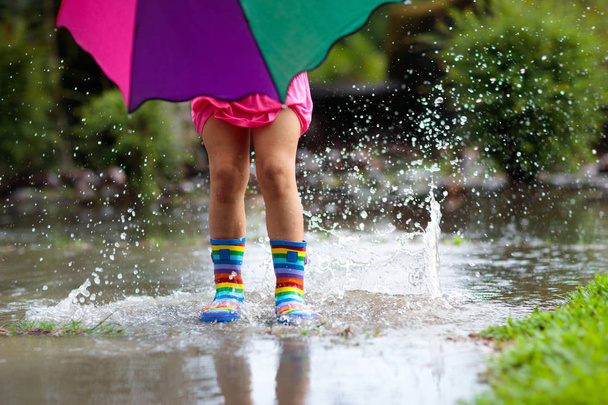 Çocuk yağmurda oyun oynuyor. Şemsiyeli ve çizmeli çocuklar sağanak yağmurda dışarıda oynuyorlar. Çamurlu su birikintisine atlayan küçük kız. Çocuklar yağmurlu sonbahar havasına göre eğlencelidir. Tropikal fırtınada koşan çocuk. - Fotoğraf, Görsel