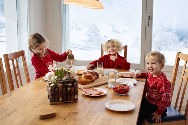 Niños desayunando en Navidad. Familia comiendo pan y bebiendo leche en casa el día de invierno nevado. Los niños comen en el comedor soleado en la ventana con montañas suizas y vista a la nieve
. - Foto, imagen