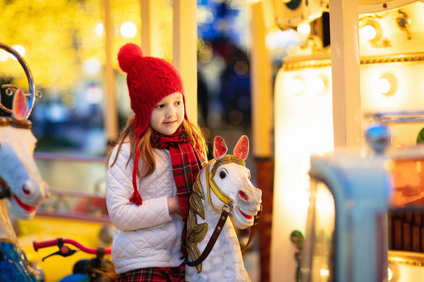 Kids at Christmas fair. Child at traditional street Xmas market in Germany. Winter outdoor fun. Little girl in knitted hat riding carousel horse in outdoor amusement park in winter holiday season.  - Photo, Image