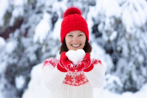 Mladá žena v pleteném svetru drží srdce ve tvaru sněhové koule v zimě. Dívka v rodinné sněhové rvačky. Žena v pleteném ručně vyrobeném klobouku a palčáky s vánočním designem sněhové vločky baví ve zasněženém parku - Fotografie, Obrázek