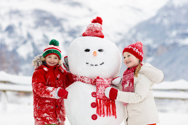 Dziecko buduje bałwana. Dzieci budują bałwana. Chłopiec i dziewczyna bawią się na dworze w śnieżny zimowy dzień. Rodzinna zabawa na świeżym powietrzu podczas świąt Bożego Narodzenia w górach. Dzieci bawią się w szwajcarskim krajobrazie górskim. - Zdjęcie, obraz
