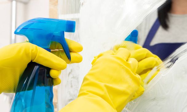 Person using spray in blue bottle to clean mirror with foam as wearing yellow rubber gloves and blue apron - Photo, image