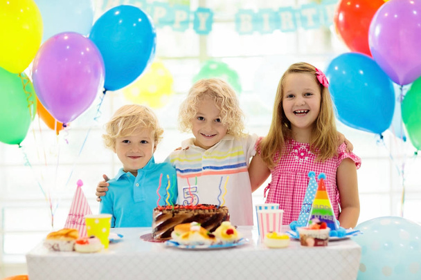 子供の誕生日パーティー。カラフルなケーキの上のろうそくを吹きの子。虹旗バナー、風船で飾られた家。ファーム動物のテーマにお祝いします。小さな少年の祝う誕生日。パーティー フード. - 写真・画像