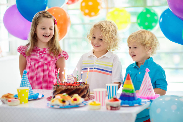 Kids birthday party. Child blowing out candles on colorful cake. Decorated home with rainbow flag banners, balloons. Farm animals theme celebration. Little boy celebrating birthday. Party food. - Foto, Imagem