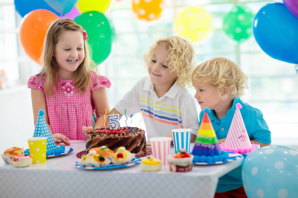 Kids birthday party. Child blowing out candles on colorful cake. Decorated home with rainbow flag banners, balloons. Farm animals theme celebration. Little boy celebrating birthday. Party food. - 写真・画像