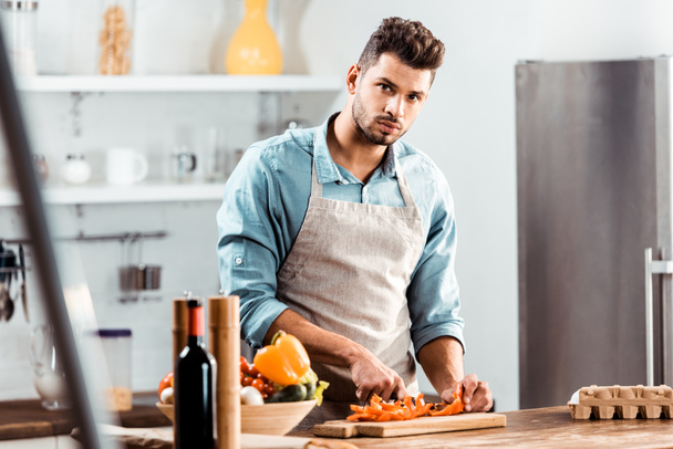 handsome young man in apron cutting vegetables and looking at camera in kitchen - Foto, afbeelding