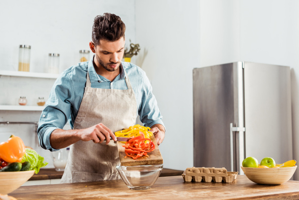 Junger Mann in Schürze bereitet Gemüsesalat in Küche zu  - Foto, Bild