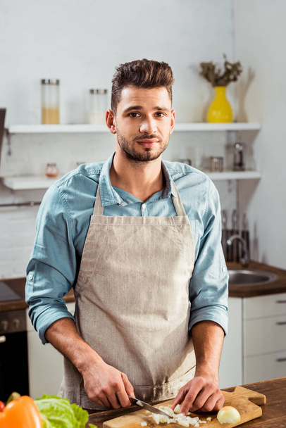 przystojny, młody człowiek w fartuch do krojenia cebuli i patrząc na kamery w kuchni - Zdjęcie, obraz