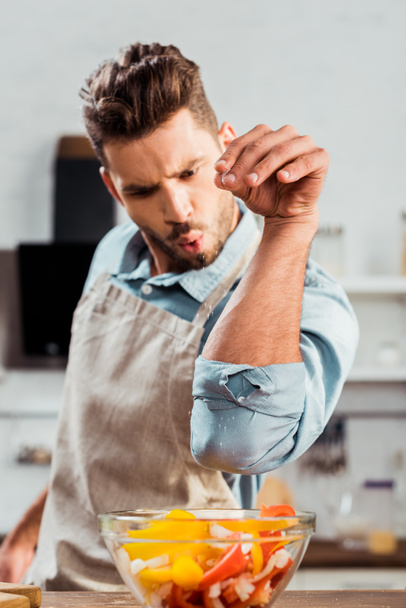 handsome young man in apron adding salt to vegetable salad  - Photo, Image