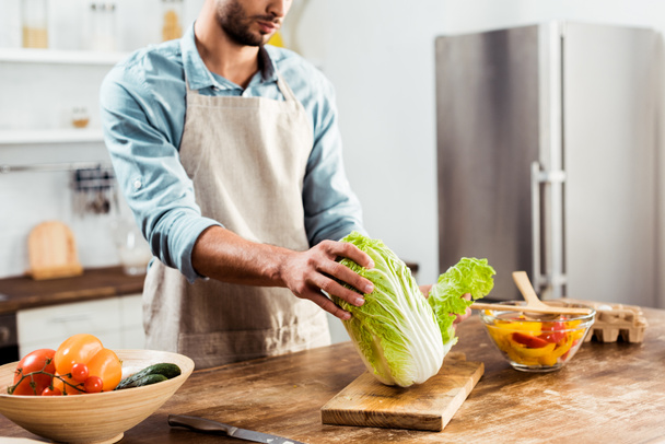 cropped shot of young man in apron holding napa cabbage and cooking in kitchen - Foto, Bild