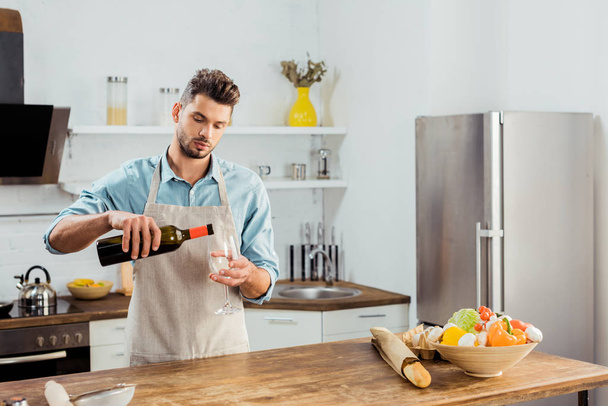 schöner junger Mann in Schürze schenkt Wein in Küche ein - Foto, Bild