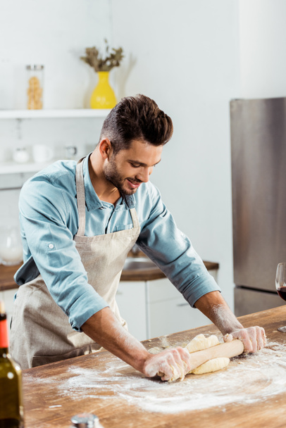 Красивый улыбающийся молодой человек в фартуке со скалкой, готовящей тесто на кухне
  - Фото, изображение