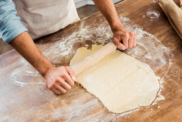 cropped shot of young man in apron holding rolling pin and preparing dough in kitchen  - Foto, afbeelding
