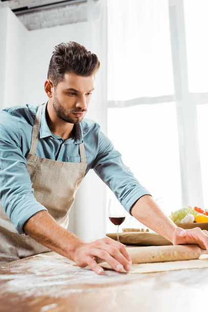 pohledný mladý muž v zástěře držící váleček a příprava těsta v kuchyni - Fotografie, Obrázek