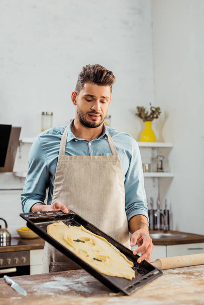 frustrated young man in apron looking at spoiled dough on baking tray - Foto, immagini
