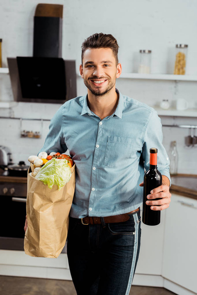handsome smiling young man holding grocery bag and bottle of wine in kitchen - Foto, Bild