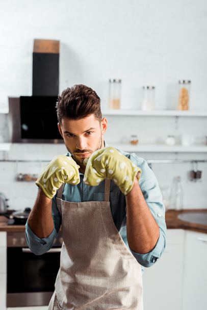 knappe jongeman in schort en pannenlappen boksen en camera kijken tijdens het koken in de keuken   - Foto, afbeelding