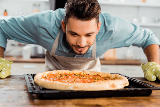 guapo sonriente joven oliendo pizza fresca casera en bandeja para hornear
 - Foto, imagen