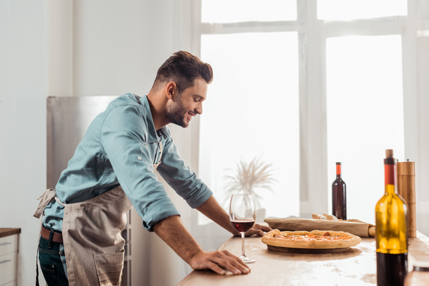 boční pohled na usmívající se mladý muž v aron dívá na čerstvé domácí pizza  - Fotografie, Obrázek